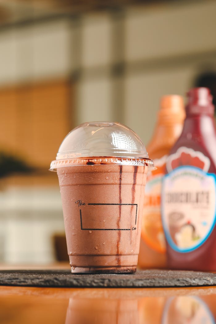Chocolate milkshake in a takeaway cup with syrup bottles in the background; perfect for food photography or menus.